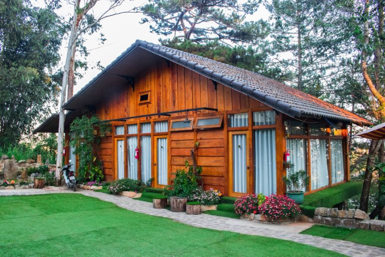 Tổng thể của Nomini homestay mang vẻ đẹp của một ngôi nhà gỗ cấp bốn, được bao quanh bởi một mảng xanh mướt của thảm cỏ và những bông hoa dại xinh xắn khoe sắc giữa tán cây thông.
